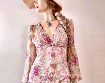 May include: A long-sleeved blouse featuring a light pink base with a vibrant floral print in shades of magenta, orange, and dark brown. The top has a deep V-neckline, gathered fabric at the shoulders, and loose sleeves, paired with blue denim jeans.