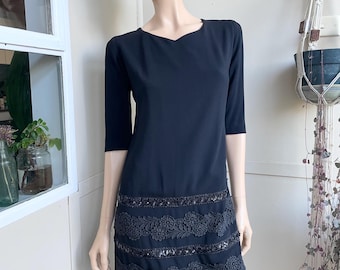 May include: A black knee-length dress featuring three-quarter length sleeves and a round neckline. The lower portion of the dress is decorated with two horizontal bands of floral lace embroidery interspersed with rows of black sequins.