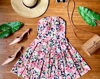 May include: A strapless floral print dress with a black background and pink, white, and green flowers. The dress has a fitted bodice and a full skirt. The dress is styled with a straw hat, sunglasses, and a wicker bag.