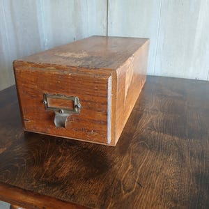 May include: A rectangular wooden storage drawer with a natural brown finish and visible wood grain. The front panel features a metallic label holder and a curved pull handle, both secured with small screws, showing signs of wear and age.
