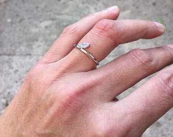 Puede incluir: Un anillo abierto de tono plateado que presenta una única piedra transparente con talla marquesa en uno de los extremos. La banda de metal fino y pulido se curva alrededor del dedo, dejando un pequeño espacio entre la piedra y el otro extremo.