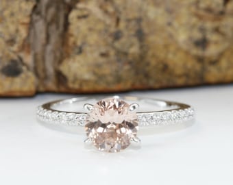 Puede incluir: Un anillo de plata con una piedra central redonda de color melocotón pálido sujeta por cuatro garras. El aro está hecho de plata pulida y está adornado con una hilera de pequeñas piedras redondas transparentes incrustadas a los lados.