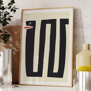 May include: A framed abstract print with a black and white design of a snake with a red mouth on a cream background.