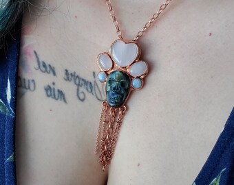 May include: A copper chain necklace with a skull pendant made of green and black stone. The pendant is surrounded by five pink and blue stones. The necklace has a tassel of copper chains.