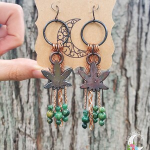 May include: A pair of dangle earrings featuring a green marijuana leaf charm, copper chain, and green beads. The earrings are displayed on a brown card with the text "Vee's Little Boutique".