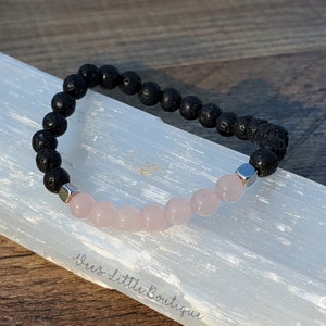 May include: A black lava rock and rose quartz beaded bracelet with silver spacer beads. The bracelet is displayed on a white selenite charging plate.