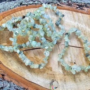 May include: A collection of green aventurine chip bracelets. The chips are irregular in shape and have a light green color with black inclusions. The bracelets are stacked on a wooden surface.