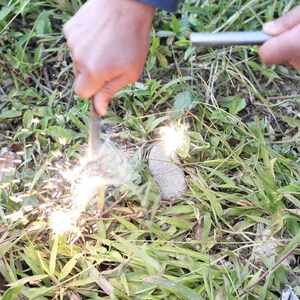 Puede incluir: Una persona est&aacute; usando un iniciador de fuego para crear chispas. El iniciador de fuego es una varilla de metal con un mango. Las chispas salen volando de la varilla y caen al suelo. El suelo est&aacute; cubierto de hierba verde.