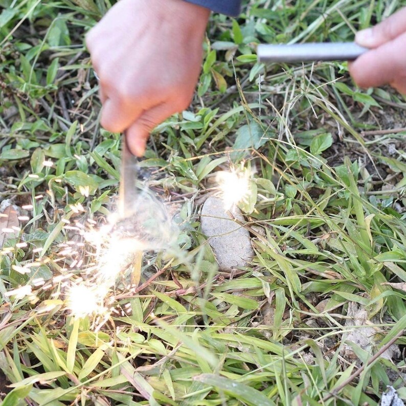 Puede incluir: Una persona est&aacute; usando un iniciador de fuego para crear chispas. El iniciador de fuego es una varilla de metal con un mango. Las chispas salen volando de la varilla y caen al suelo. El suelo est&aacute; cubierto de hierba verde.
