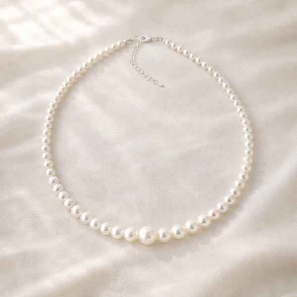 May include: A delicate pearl necklace with a graduated design. The necklace features small, round, white pearls that increase in size towards the center, with a larger pearl as the focal point. The necklace has a silver-colored clasp and extender chain.