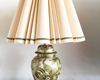 May include: A table lamp with a white ceramic base decorated with green and gold peacocks. The lamp has a pleated, off-white lampshade with green trim.