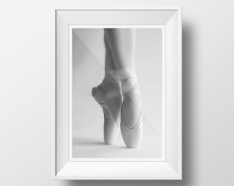 May include: A black and white photograph of a ballerina's feet wearing white pointe shoes with white ribbons. The photo is framed in a white frame.
