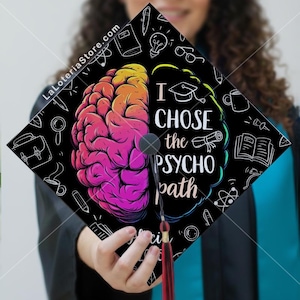 May include: Black graduation cap with a colorful brain graphic and the text "I CHOSE the PSYCHO path." The design includes academic icons and a red tassel. The cap is held by a person.