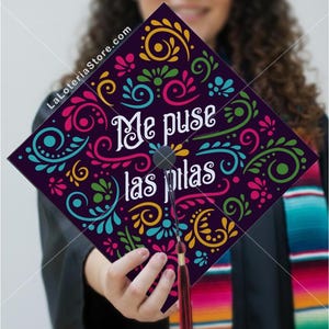 May include: A graduation cap with a dark purple background and colorful floral designs in pink, yellow, green, and teal. The text "Me puse las pilas" is written in white. A red tassel hangs from the cap.