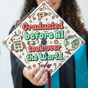 May include: A graduation cap with the text "Graduated before AI took over the World." The design includes a robot, a globe, and other graphics. The cap has a red tassel and the name "Taylor" and the year "26" are also visible.