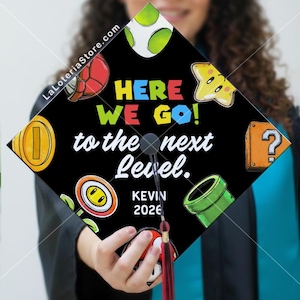 May include: Black graduation cap decorated with colorful Super Mario Bros. themed graphics. The cap features the text "HERE WE GO! to the next Level." and the name "KEVIN 2026". Includes a red tassel.