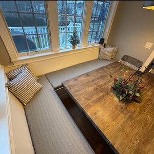 May include: A wooden dining table with a dark brown finish and a black metal base. The table is set with a centerpiece of greenery and red berries. The table is surrounded by a built-in bench with gray cushions and striped pillows.
