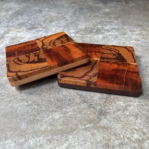 May include: Two rectangular wooden coasters with a patchwork design. The coasters feature a mix of reddish-brown and light brown wood grains, creating a visually appealing contrast. The edges are smooth and finished, showcasing the natural wood patterns.