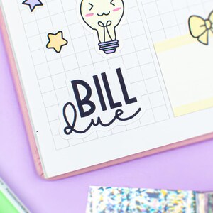 Op de afbeelding: Een planner met stickers, een groene pen en glittertape op een paars oppervlak. De planner heeft een rasterpatroon en stickers met sterren, een gloeilamp en een strik. Het woord "BILL" staat op een sticker.