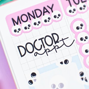 May include: A planner page with pink headers for Monday and Tuesday, each adorned with skull graphics. The text "DOCTOR appt" is visible. A colorful bookmark and a decorative pen are in the background.