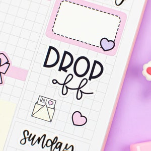 Könnte beinhalten: Eine Planerseite mit Aufklebern und einem Stift. Die Aufkleber enthalten Herzen, Schleifen und einen Kuchen. Auf den Aufklebern steht "Friday Important", "Saturday", "Drop Off" und "Sunday". Ein weißer und rosa Stift mit einem bärenförmigen Radiergummi befindet sich in der Ecke.