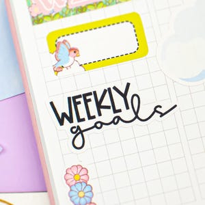 Può includere: Un'agenda rosa e bianca con adesivi decorativi. Gli adesivi includono disegni floreali, una nuvola e un uccello. Le parole "WEEKLY goals" sono scritte in corsivo nero. L'agenda è aperta su una pagina a griglia.