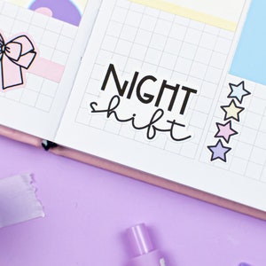 May include: A close-up of a planner with a sticker that says "NIGHT shift" and a row of colorful star stickers. The planner is open to a page with a grid pattern.