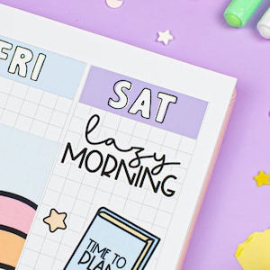 May include: A weekly planner with a grid layout and colorful stickers. The stickers include a rainbow, a star, a bow, and a book with the text "Time to Plan". The planner has the days of the week labeled, including "FRI" and "SAT".