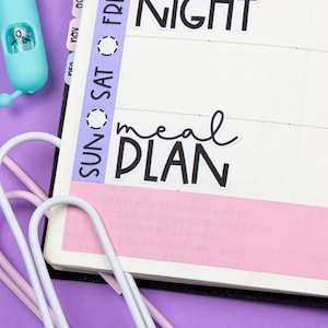 May include: A close-up of a planner with a pink cover and a sticker that says "meal plan". The planner has a tab that says "SUN SAT FRI" and another tab that says "NIGHT". There are also rainbow heart stickers that spell out "WEEKEND".