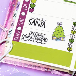 Op de afbeelding: Een plannerpagina met stickers met een vakantiethema. De stickers bevatten een kerstboom, cadeauverpakkingen en de zinnen "visit SANTA" en "DECORATE GINGERBREAD house". De planner heeft een paarse en roze kaft met washi tape.