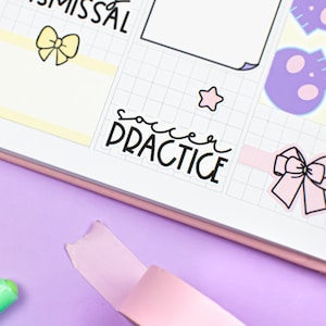 May include: A pink washi tape dispenser with a roll of pink washi tape. The dispenser is on a purple surface with colorful markers and a planner with stickers that say "Dismissal" and "Soccer Practice".
