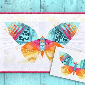 Könnte beinhalten: Ein farbenfroher Schmetterlings-Quiltblock mit rosa Rand. Der Schmetterling besteht aus verschiedenen Stoffen in Orange-, Gelb-, Türkis- und Rosatönen. Der Quiltblock befindet sich auf einem hellblauen Holzgrund.