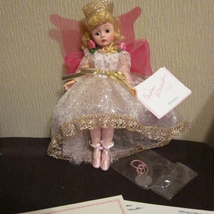 May include: A porcelain doll wearing a white dress with gold trim and a pink cape. The doll has blonde hair and is wearing a gold crown. The doll is sitting on a white background. The doll is part of the Madame Alexander collection and has the number 159130.