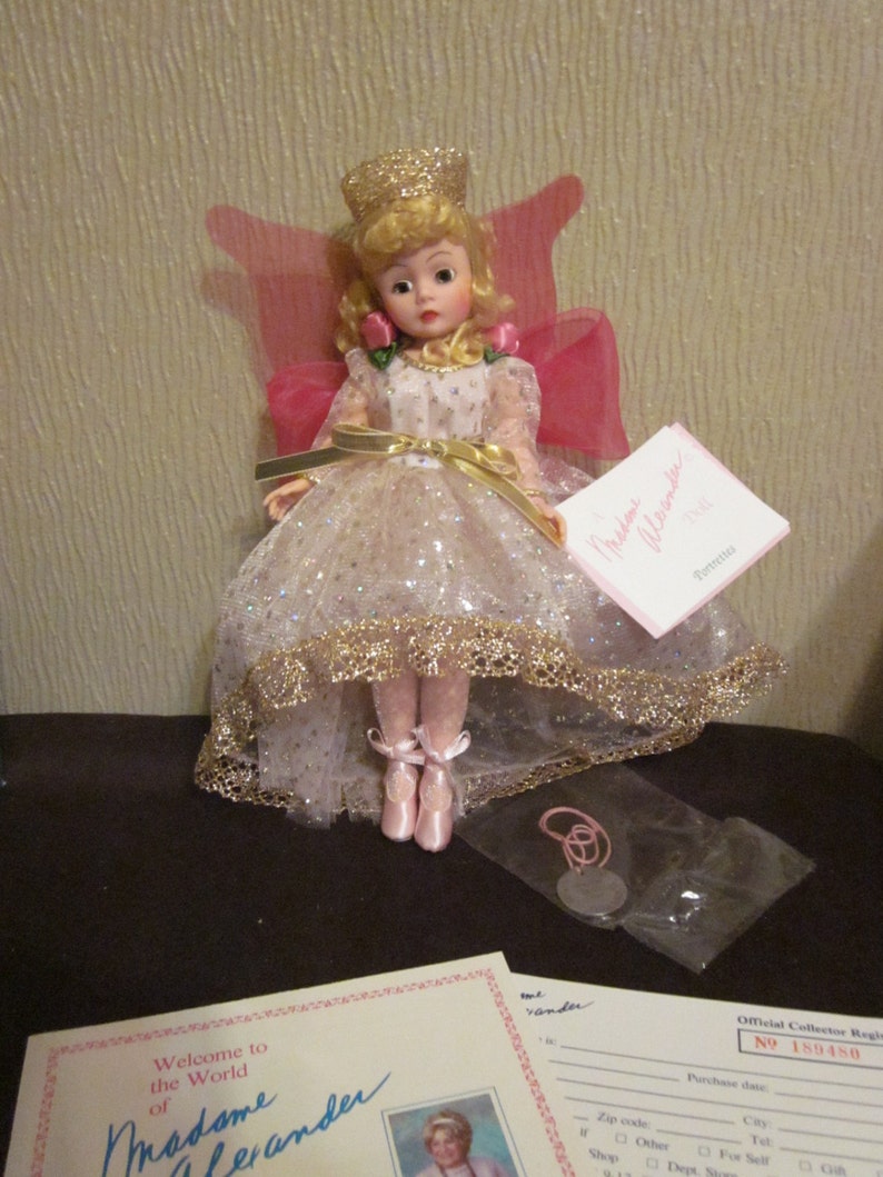 May include: A porcelain doll wearing a white dress with gold trim and a pink cape. The doll has blonde hair and is wearing a gold crown. The doll is sitting on a white background. The doll is part of the Madame Alexander collection and has the number 159130.