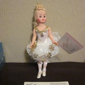 K&ouml;nnte beinhalten: Eine Porzellanpuppe in einem wei&szlig;en und goldenen Ballerina-Kost&uuml;m mit einem Schneeflockenmuster. Die Puppe hat blonde Haare und blaue Augen. Die Puppe h&auml;lt eine kleine wei&szlig;e und goldene Schneeflocke in ihrer rechten Hand. Die Puppe steht auf einer braunen Oberfl&auml;che. Die Puppe tr&auml;gt wei&szlig;e Ballettschuhe. Die Puppe h&auml;lt eine Karte mit der Aufschrift "Madame Alexander".