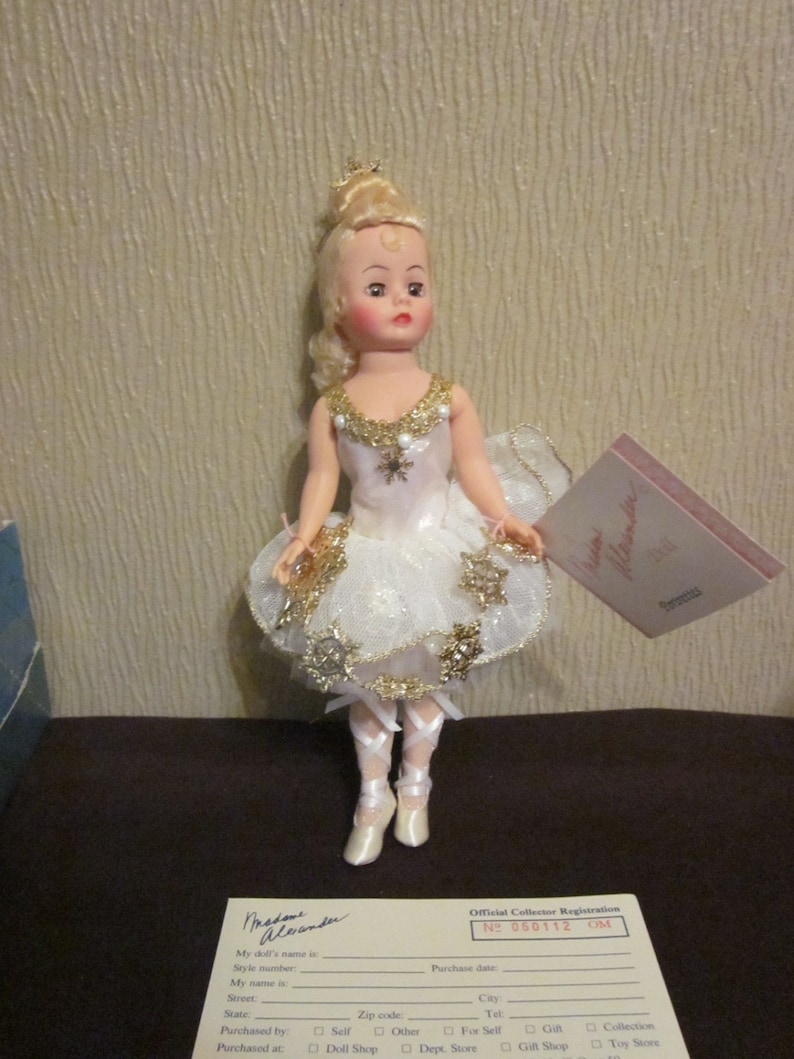 K&ouml;nnte beinhalten: Eine Porzellanpuppe in einem wei&szlig;en und goldenen Ballerina-Kost&uuml;m mit einem Schneeflockenmuster. Die Puppe hat blonde Haare und blaue Augen. Die Puppe h&auml;lt eine kleine wei&szlig;e und goldene Schneeflocke in ihrer rechten Hand. Die Puppe steht auf einer braunen Oberfl&auml;che. Die Puppe tr&auml;gt wei&szlig;e Ballettschuhe. Die Puppe h&auml;lt eine Karte mit der Aufschrift "Madame Alexander".