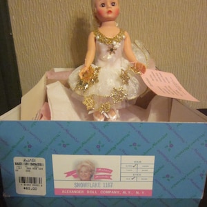 K&ouml;nnte beinhalten: Eine wei&szlig;e und goldene Ballerina-Puppe mit blonden Haaren und blauen Augen. Die Puppe tr&auml;gt ein wei&szlig;es T&uuml;llkleid mit goldenen Schneeflocken und einer goldenen Krone. Die Puppe befindet sich in einer Schachtel mit den Worten "Snowflake 1167" und "Alexander Doll Company, N.Y., N.Y." darauf.
