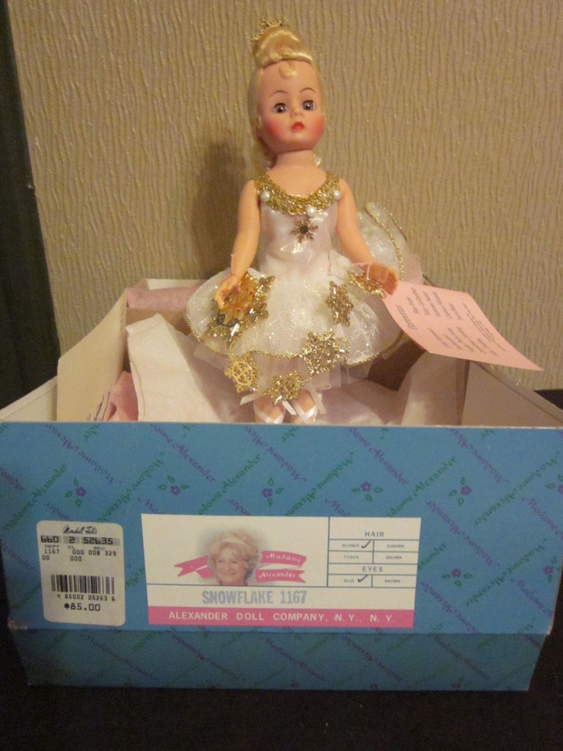 K&ouml;nnte beinhalten: Eine wei&szlig;e und goldene Ballerina-Puppe mit blonden Haaren und blauen Augen. Die Puppe tr&auml;gt ein wei&szlig;es T&uuml;llkleid mit goldenen Schneeflocken und einer goldenen Krone. Die Puppe befindet sich in einer Schachtel mit den Worten "Snowflake 1167" und "Alexander Doll Company, N.Y., N.Y." darauf.