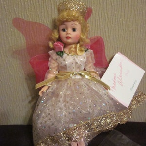 May include: A porcelain doll with blonde hair and a pink and gold dress. The doll is wearing a crown and has wings. The doll is holding a pink rose. The doll is sitting on a black surface. The text on the card reads "Madame Alexander Doll".