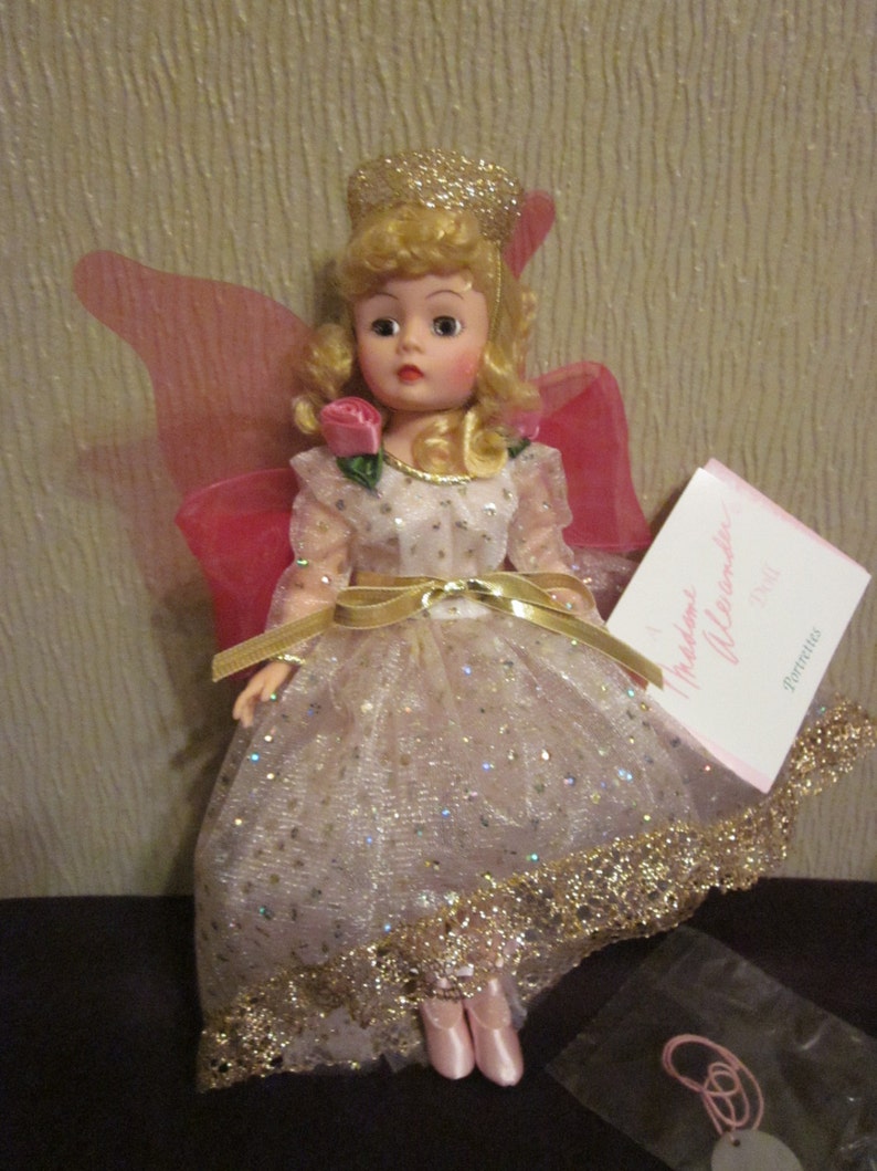 May include: A porcelain doll with blonde hair and a pink and gold dress. The doll is wearing a crown and has wings. The doll is holding a pink rose. The doll is sitting on a black surface. The text on the card reads "Madame Alexander Doll".