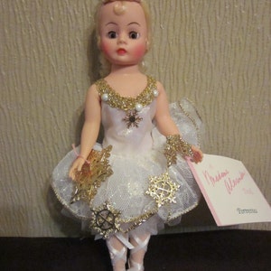K&ouml;nnte beinhalten: Eine Porzellanpuppe, die als Ballerina verkleidet ist und ein wei&szlig;es Tutu mit goldenem Glitzer und Schneeflocken-Akzenten tr&auml;gt. Die Puppe hat blonde Haare, blaue Augen und rosige Wangen. Die Puppe h&auml;lt eine goldene Schneeflocke in der Hand. Die Puppe ist mit "Madame Alexander Doll" beschriftet.