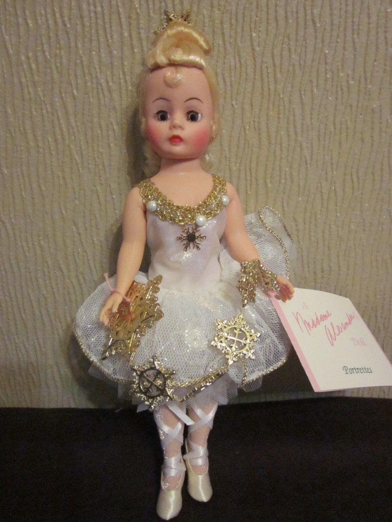 K&ouml;nnte beinhalten: Eine Porzellanpuppe, die als Ballerina verkleidet ist und ein wei&szlig;es Tutu mit goldenem Glitzer und Schneeflocken-Akzenten tr&auml;gt. Die Puppe hat blonde Haare, blaue Augen und rosige Wangen. Die Puppe h&auml;lt eine goldene Schneeflocke in der Hand. Die Puppe ist mit "Madame Alexander Doll" beschriftet.
