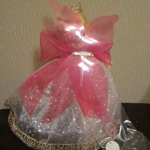 May include: A pink and white fairy doll with a gold crown and wings. The doll is wearing a pink and white dress with gold trim and a pink bow.