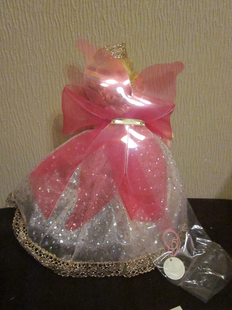 May include: A pink and white fairy doll with a gold crown and wings. The doll is wearing a pink and white dress with gold trim and a pink bow.
