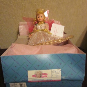 May include: A tooth fairy doll wearing a pink and gold dress with a crown and wings. The doll is sitting in a pink box with a blue label that says "Tooth Fairy 201560" and "Alexander Doll Company, NY, NY".