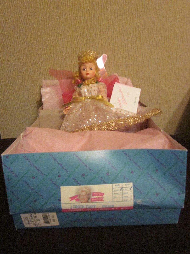 May include: A tooth fairy doll wearing a pink and gold dress with a crown and wings. The doll is sitting in a pink box with a blue label that says "Tooth Fairy 201560" and "Alexander Doll Company, NY, NY".