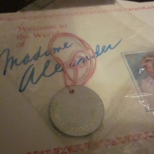 May include: A pink string attached to a silver round tag with the text "The Doll's Own" on it. The tag is on a white card with the text "Welcome to the World of Madame Alexander" in blue ink.