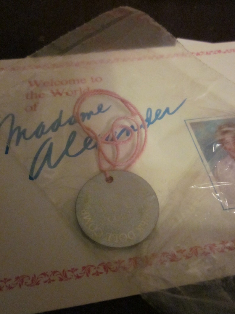 May include: A pink string attached to a silver round tag with the text "The Doll's Own" on it. The tag is on a white card with the text "Welcome to the World of Madame Alexander" in blue ink.