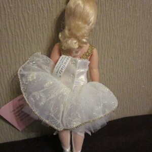 K&ouml;nnte beinhalten: Eine Porzellanpuppe, die als Ballerina verkleidet ist, tr&auml;gt ein wei&szlig;es T&uuml;ll-Tutu mit goldenen Verzierungen und eine goldene Krone. Die Puppe hat blonde Haare und steht auf einer schwarzen Oberfl&auml;che. Die Puppe ist mit "Snowflake" und "Little Memories by Ashton Drake Galleries" beschriftet.