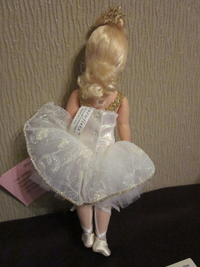 K&ouml;nnte beinhalten: Eine Porzellanpuppe, die als Ballerina verkleidet ist, tr&auml;gt ein wei&szlig;es T&uuml;ll-Tutu mit goldenen Verzierungen und eine goldene Krone. Die Puppe hat blonde Haare und steht auf einer schwarzen Oberfl&auml;che. Die Puppe ist mit "Snowflake" und "Little Memories by Ashton Drake Galleries" beschriftet.